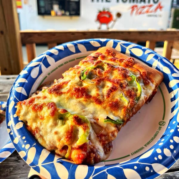 A slice of The Tomato Pizza's cheesy pizza with green peppers on a blue and white paper plate.