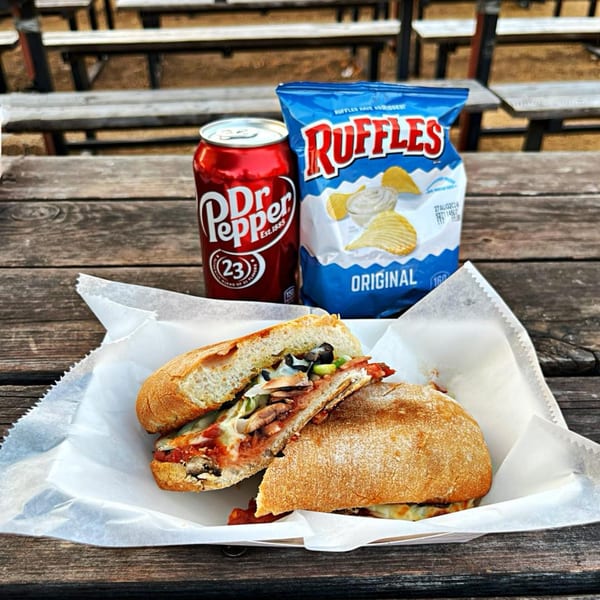 A pizza sub, Dr. Pepper, and Ruffles chips on a rustic wooden picnic table.