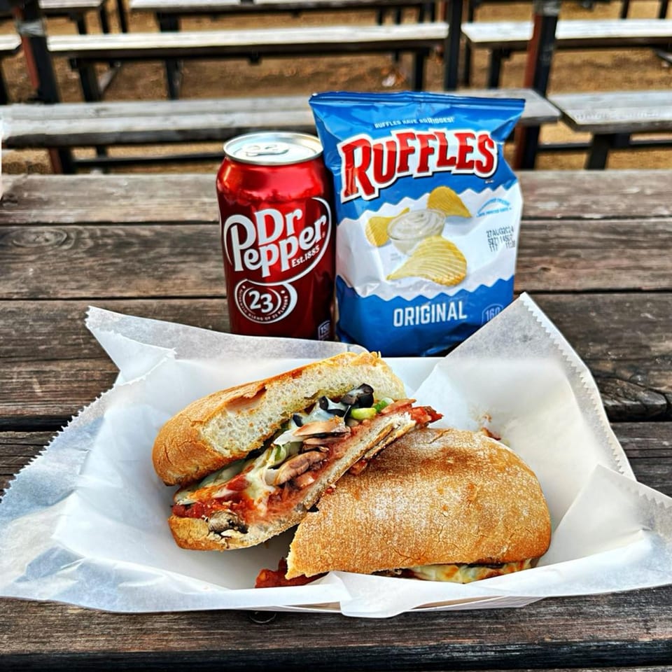 A pizza sub, Dr. Pepper, and Ruffles chips on a rustic wooden picnic table.
