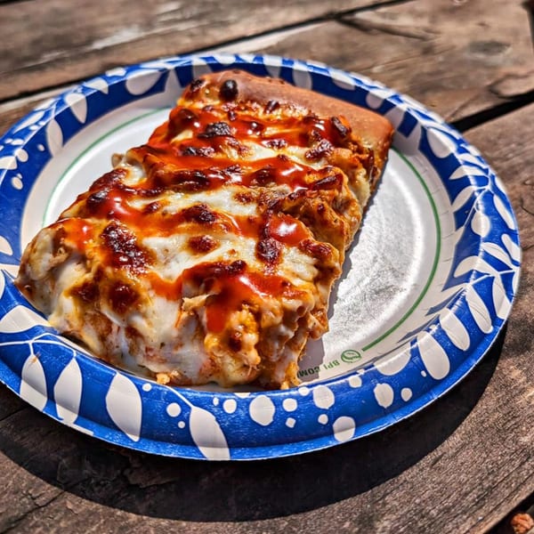 A slice of The Tomato Pizza's buffalo chicken pizza on a paper plate on a wooden table.