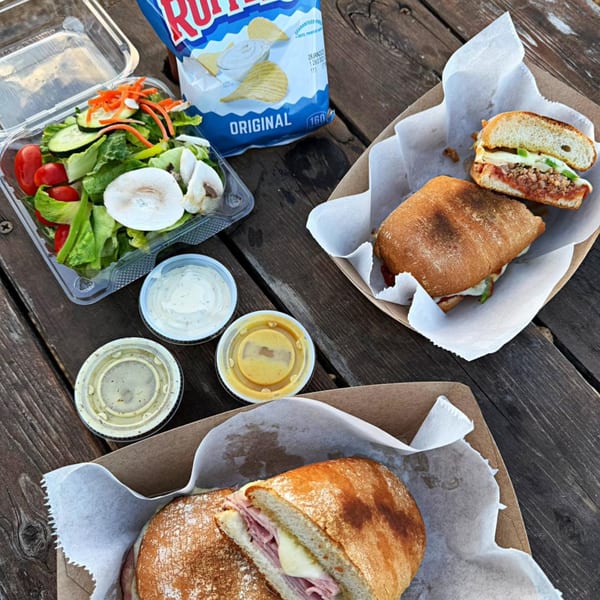 Two sandwiches, a side salad, chips, and three dipping sauces on a rustic wooden table.