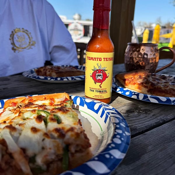Slices of pizza on paper plates with a bottle of "Tomato Tears" hot sauce on a wooden table.